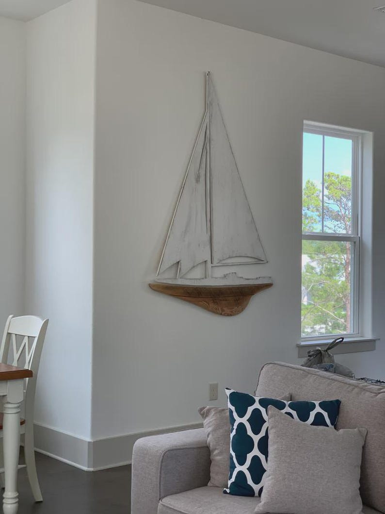 May include: A white sailboat wall decor with a wooden base is mounted on a white wall. The sailboat has a simple design with a mast and sail. The room also features a gray sofa and decorative pillows.