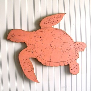 May include: A coral pink wooden sea turtle wall decor with a detailed shell and flippers. The turtle is painted in a distressed style, giving it a vintage look.