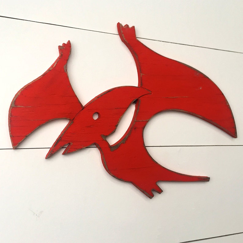 May include: Red wooden cutout of a pterodactyl, a flying dinosaur with a long beak and wings. The cutout is painted a bright red color and has a distressed finish.