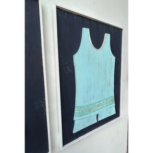 Pu&ograve; includere: Un'opera d'arte in legno incorniciata con un design di costume da bagno vintage azzurro su sfondo blu scuro. Il costume da bagno ha un dettaglio a righe decorative. L'opera d'arte &egrave; racchiusa in una cornice bianca.