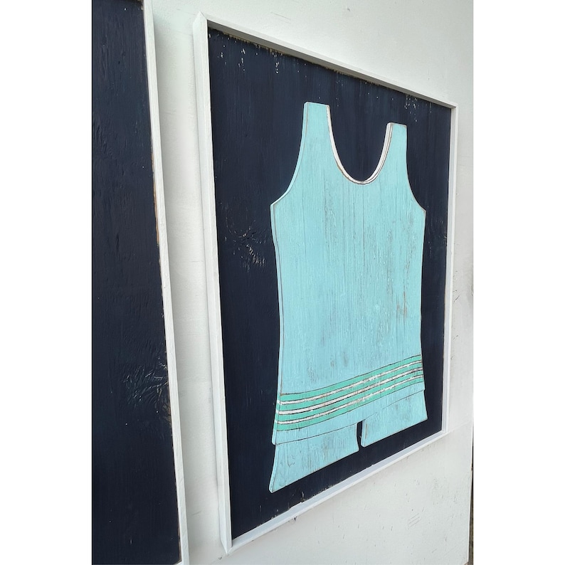 Pu&ograve; includere: Un'opera d'arte in legno incorniciata con un design di costume da bagno vintage azzurro su sfondo blu scuro. Il costume da bagno ha un dettaglio a righe decorative. L'opera d'arte &egrave; racchiusa in una cornice bianca.