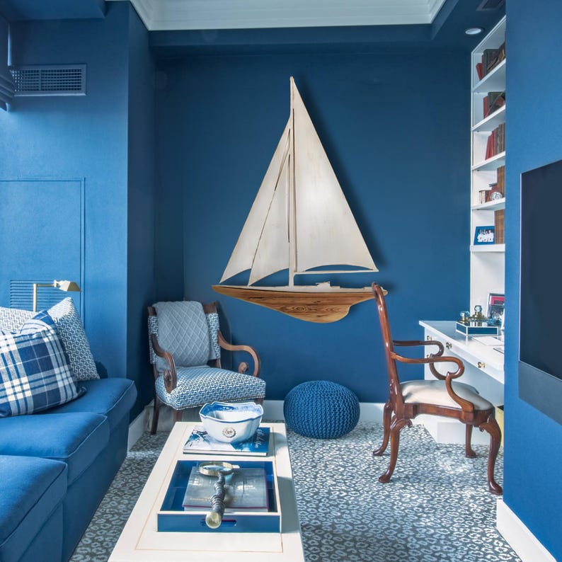 May include: A nautical-themed room with blue walls and furniture. A wooden sailboat wall decor is mounted above a chair. A white coffee table with a blue tray and a blue pouf add to the coastal aesthetic.