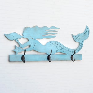 Puede incluir: Un gancho de pared de madera de color azul claro en forma de sirena con tres ganchos de metal negro. La sirena sostiene una concha y una estrella de mar.