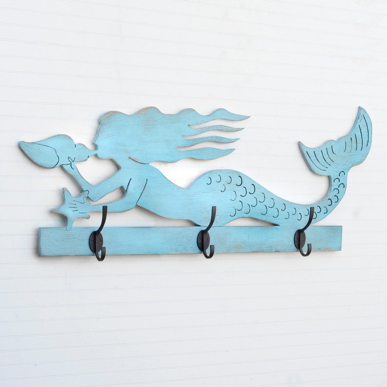 Puede incluir: Un gancho de pared de madera de color azul claro en forma de sirena con tres ganchos de metal negro. La sirena sostiene una concha y una estrella de mar.