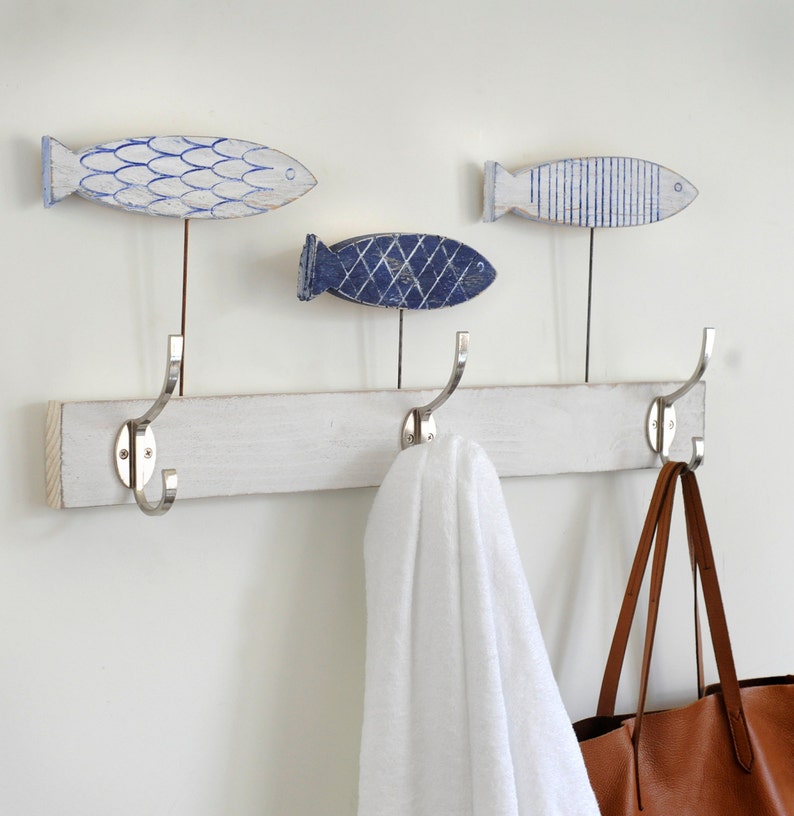 May include: White wooden coat rack with three hooks and three blue and white fish-shaped decorations. A white towel hangs on one hook and a brown leather tote bag hangs on another hook.