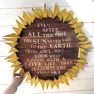 Könnte beinhalten: Ein rundes Holzschild mit einem Sonnenblumen-Design. Die Mitte besteht aus braunen Brettern mit weißem Text, der lautet: „AUCH NACH ALL DIESER ZEIT SAGT DIE SONNE NIEMALS ZUR ERDE: 'DU SCHULDEST MIR ETWAS.' SIEH, WAS MIT LIEBE WIE DIESER GESCHIEHT, SIE ERHELLT DEN GANZEN HIMMEL.“
