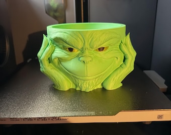 May include: A lime green planter shaped like the Grinch's face, with his hands supporting his cheeks. The planter has a wide opening and is sitting on a black surface. The planter is approximately 10 inches wide.