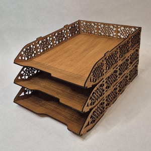 Könnte beinhalten: Ein dreistufiger Schreibtisch-Organizer aus Holz mit hellbraunem Finish. Jedes Fach hat ein dekoratives, durchbrochenes Design an den Kanten. Der Organizer ist für die Aufnahme von Dokumenten und Papieren konzipiert und bietet eine stilvolle Aufbewahrungslösung.