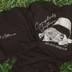 May include: Two dark brown t-shirts on green grass. One shirt has the text "Abnormal Attire co." in white script. The other shirt features a raccoon graphic with the text "Complexly Southern" and "We're just as confused as you."