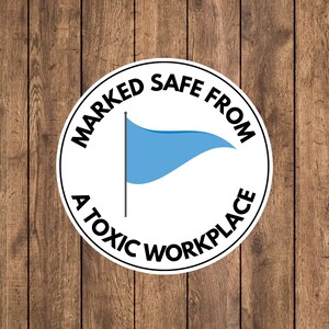 May include: A round white sticker with black text and a light blue flag graphic. The text reads "MARKED SAFE FROM A TOXIC WORKPLACE" in a circular arrangement around the flag. The sticker is on a wooden background.