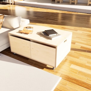 May include: A light-colored wooden coffee table with two drawers and a rectangular shape. The table has a smooth surface and is on wheels. Two notebooks and a small wooden object are on top of the table.