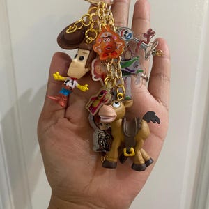 May include: A collection of Toy Story character keychains on a gold chain. The keychains include Woody, Buzz Lightyear, Mr. Potato Head, and others. The keychains are made of plastic and metal, and are attached to a gold-colored keyring.