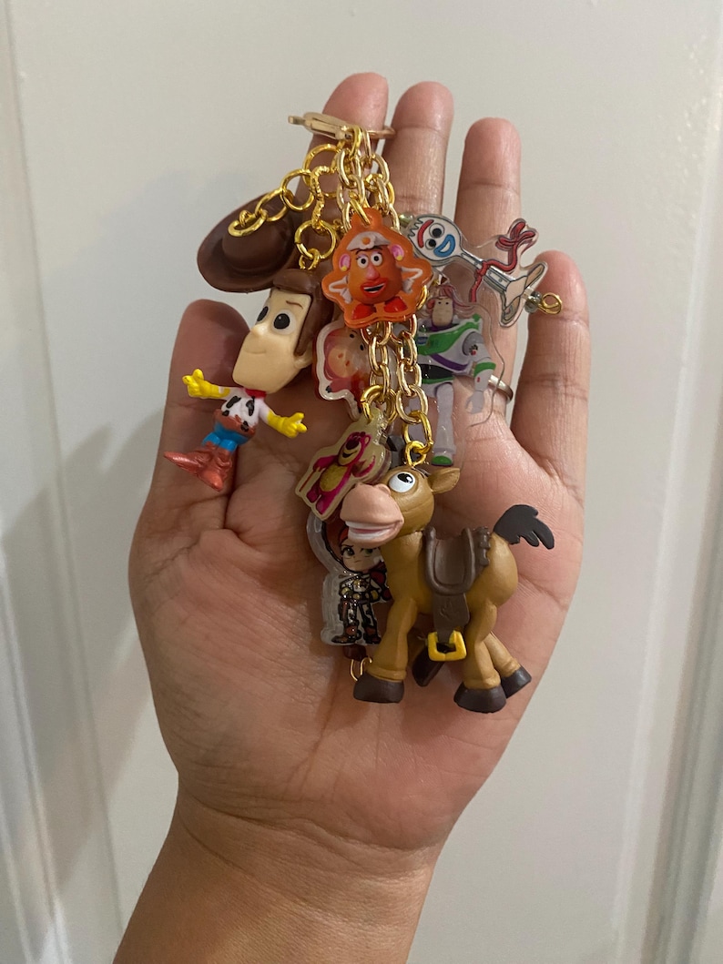 May include: A collection of Toy Story character keychains on a gold chain. The keychains include Woody, Buzz Lightyear, Mr. Potato Head, and others. The keychains are made of plastic and metal, and are attached to a gold-colored keyring.