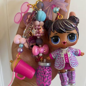 May include: A colorful keychain featuring an LOL doll, pink sunglasses, a pink sippy cup, and a jar with a leopard print design. The doll has dark hair and is wearing a pink and blue leopard print jacket and boots. The keychain includes various beads and charms.
