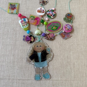 May include: A decorative wall hanging featuring a large central Cabbage Patch Kids acrylic charm of a blonde doll in a blue dress and black jacket, surrounded by various smaller colorful charms, beads, and printed Cabbage Patch Kids logos.