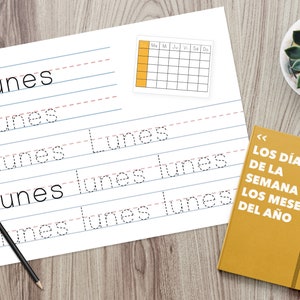 Puede incluir: Una hoja de papel blanca con l&iacute;neas de escritura para practicar la escritura de la palabra espa&ntilde;ola "Lunes" (lunes). El papel est&aacute; sobre una superficie de madera con un calendario amarillo y un libro amarillo con el texto "LOS D&Iacute;AS DE LA SEMANA Y LOS MESES DEL A&Ntilde;O" (Los d&iacute;as de la semana y los meses del a&ntilde;o).