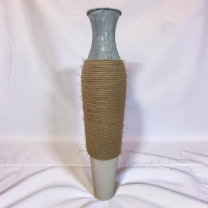 May include: A tall, slender vase featuring a flared top section with a distressed gray finish. The central body is wrapped in thick, natural-colored jute rope with visible frayed fibers, while the base is a smooth, solid light gray cylindrical form.