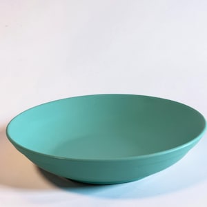 May include: A solid turquoise-colored bowl with a wide, shallow design. The bowl has a smooth, matte finish and a simple, minimalist aesthetic. The bowl is suitable for serving salads, pasta, or other dishes.