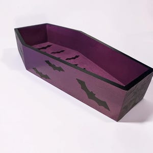 May include: A coffin-shaped tray with a deep purple interior and exterior. The exterior sides are decorated with black bat silhouettes. The top edges of the tray are finished with a solid black border, creating a distinct contrast against the purple.