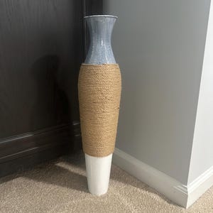 May include: A tall decorative vase with a gray top, a tan rope-wrapped midsection, and a white base. The vase is cylindrical and stands on a beige carpet, with a dark brown door and a gray wall in the background.