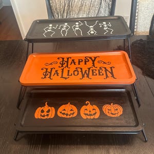 May include: A three-tiered rectangular display stand with black metal legs. The top tray is black with white dancing skeletons. The middle tray is orange with black text reading Happy Halloween. The bottom tray is black with orange jack-o'-lanterns.