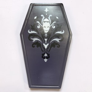 May include: A coffin-shaped wall plaque painted in a dark purple hue. The center features a detailed damask-style pattern in black and white, incorporating a central skull motif with intricate swirling flourishes extending upwards and downwards.