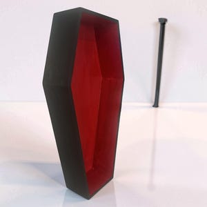 May include: A coffin-shaped box with a matte black exterior and a vibrant red interior. The box is open, revealing the hollow red space inside. A separate, thin, black, rod-like piece stands vertically in the background, detached from the main box.