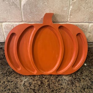 May include: Orange wooden pumpkin-shaped serving tray with a stem. The tray has a central oval section and three curved sections on each side. The tray is painted in a solid orange colour and is designed for serving food or displaying items.