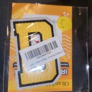 May include: A yellow embroidered letter D patch with a black border and white trim, enclosed in a clear plastic bag. A white barcode label is attached to the front, displaying the text: X002MAEPGR Letter D Iron on Patc... 16 inches (Yellow, D).