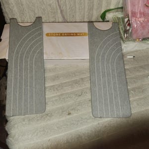 May include: Two rectangular grey stone drying mats featuring a textured surface with a series of curved, arched lines. Each mat has a semi-circular cutout at the top. Behind the mats is a white product box labeled with the text STONE DRYING MAT.