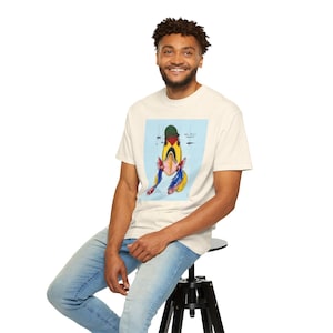May include: Cream-colored t-shirt featuring a colorful fish illustration on a light blue background. The fish has a yellow body, red and blue fins, and a green hat. The shirt is worn by a person sitting on a black stool.