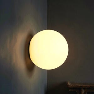 May include: A round, glowing, white wall-mounted light fixture. The light is emitting a soft, warm glow against a dark gray wall. The fixture is mounted in the corner of a room.