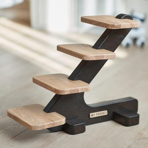 May include: A wooden pet step with four platforms. The steps are made of light-colored wood and are attached to a dark brown frame. The base of the step has the text "MY FRIEND" on it. The steps are designed to help pets reach higher places.