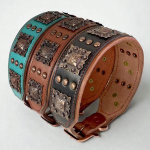 May include: A stack of wide dog collars in teal, brown, and black leather. Each collar is adorned with copper-colored square embellishments and rivets. The collars have a buckle closure and are designed for durability and style.