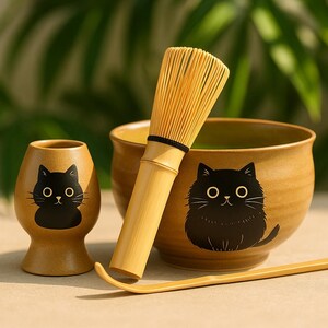 Puede incluir: Un juego de té matcha con un batidor de bambú, un cuenco de cerámica, una taza de cerámica pequeña y una cuchara de bambú. Cada pieza de cerámica tiene una ilustración de un gato negro. El juego es en tonos beige y marrón.