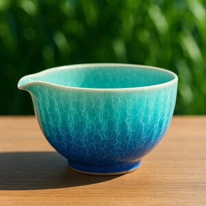 May include: A ceramic tea bowl with a turquoise and blue gradient glaze. The bowl has a spout and a crackle pattern, sitting on a wooden surface. The background is blurred green.
