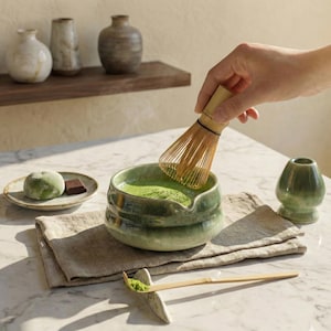 Könnte beinhalten: Matcha-Tee wird in einer grünen Keramikschale mit einem Bambusbesen verrührt. Ein kleiner Teller mit einem grünen Mochi und einem Stück dunkler Schokolade befindet sich in der Nähe. Eine kleine grüne Keramikvase ist ebenfalls vorhanden.
