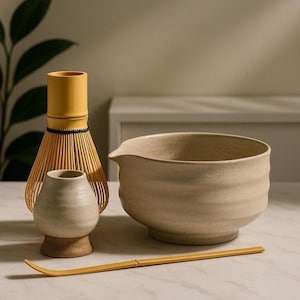 Op de afbeelding: Een matcha-theeset met een gele bamboe klopper, een kleine keramische theekom, een beige keramische kom met een tuit en een gele bamboelepel. De set is voor het bereiden van matcha-thee.