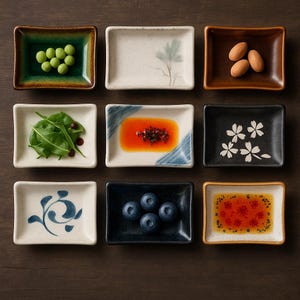 Può includere: Nove piccoli piatti rettangolari in ceramica disposti su una superficie di legno scuro. Ogni piatto contiene un alimento diverso, tra cui piselli verdi, mandorle, mirtilli e salse. Alcuni piatti hanno motivi floreali decorativi.