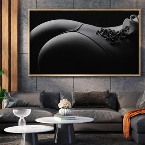 Puede incluir: Impresión artística en blanco y negro que muestra la espalda de una persona con una taza de café y granos. La imagen está enmarcada en un marco dorado y cuelga sobre un sofá moderno.