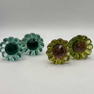 May include: Four decorative glass cabinet knobs. Two are a light teal color with a dark green center. The other two are yellow-green with a brown center. The knobs are shaped like flowers.