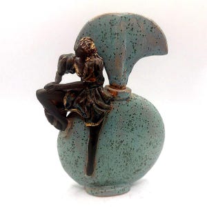 Pode incluir: Uma escultura de cerâmica azul-turquesa de uma mulher sentada numa base redonda. A mulher é castanha escura com detalhes dourados, vestindo um vestido. A escultura tem um elemento em forma de leque atrás da mulher.