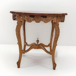May include: A light brown wooden console table featuring a semi-oval top and an ornate carved apron with shell motifs. The table stands on four curved legs connected by a central stretcher that supports a small, decorative finial in the center.