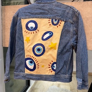 Pode incluir: Um casaco de ganga azul com um grande painel retangular bege claro nas costas. O painel é decorado com motivos de olhos azuis e brancos pintados à mão, linhas radiantes castanhas e pequenas estrelas amarelas espalhadas pelo tecido.