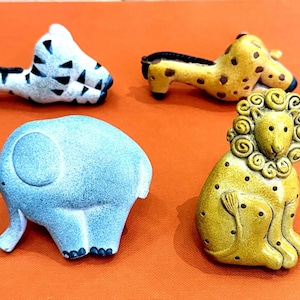 May include: Four animal figurines including a white and black zebra, a yellow and brown giraffe, a light blue elephant, and a yellow lion with a textured mane and black spots. Each piece is crafted with a smooth finish and distinct features.