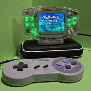 May include: A clear Game Boy Advance handheld console with glowing green internal lights displaying the Pokémon Emerald Version title screen, resting on a black carrying case behind a grey Super Nintendo controller with purple buttons.