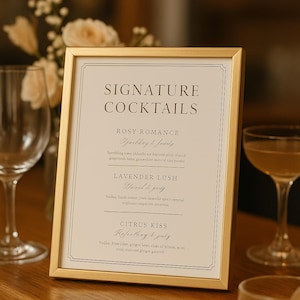 May include: A gold-framed sign with the words "SIGNATURE COCKTAILS" in elegant lettering. Below, three cocktail names are listed: Rosy Romance, Lavender Lush, and Citrus Kiss. The sign is on a wooden surface.