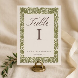 May include: A wedding table card with a floral border in olive green. The card reads "Table I" and includes the names "Gwynth & Gordon" and the date "11.02.2030". The card is held by a gold-colored metal holder.