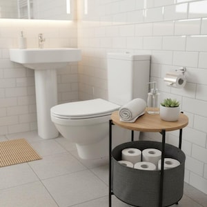 May include: A contemporary bathroom interior with a white toilet, a pedestal sink, and a small round side table. The table has a wooden top and a grey fabric basket for toilet rolls. A rolled towel and a small plant are on the table.