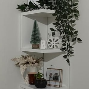 Op de afbeelding: Een witte hoekplank met diverse decoratieve items. Inclusief een gouden draadmand, ingelijste kunst, een kleine kerstboom, een keramische sneeuwvlok, een vaas met gedroogde bloemen en een vintage radio.